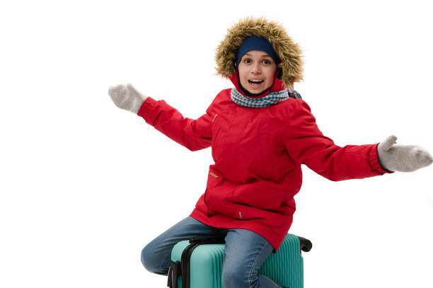 Cheerful boy wearing red parka, cutely smiling to camera, sitting on blue suitcase, traveling abroad at weekends getaway Cheerful boy in red parka with fur hood and woolen mittens, smiling with toothy smile, looking at camera, sitting on blue suitcase, traveling abroad on weekends or winter holidays, isolated on white cutely stock pictures, royalty-free photos & images