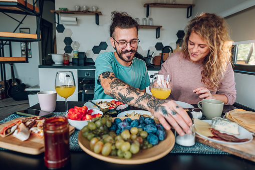 Beautiful happy couple eating breakfast together while sitting at table at home. Focus on a tattooed man with eyeglasses. Romantic spouses eating tasty food And chatting. Healthy lifestyle.