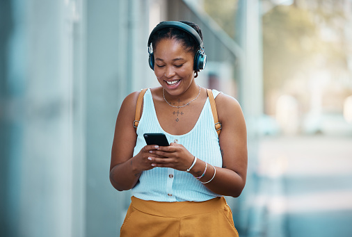 Urban black woman with smartphone and headphones in city or street walking and listening to music, networking and typing on chat ap. Gen z girl with cellphone with subscription or 5g audio streaming