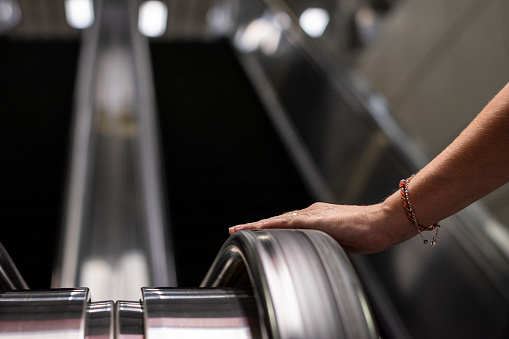 Person about to climb a large escalator. Person at the base of an escalator. Hand of a person holding on to the handrail of a staircase. Inside the subway station.