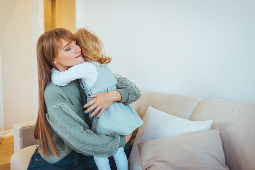 Lovely mother embracing her cute daughter on the sofa at home. Lovely Family. Mother embracing her daughter, feeling happy and carefree. Copy space. Loving young mother laughing embracing smiling cute funny kid girl