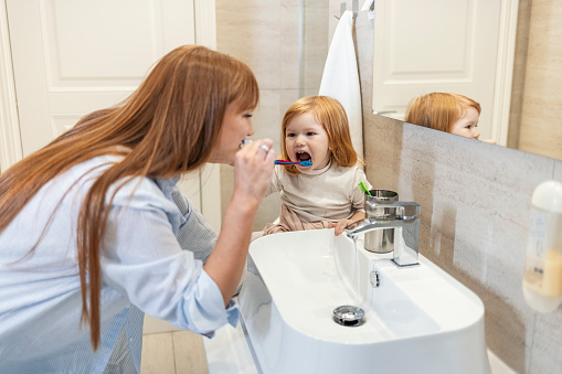 Mother watching daughter brushing teeth at home. Happy woman brushing teeth with her daughter. Little girl brushing teeth with her mom. Smiling mother and daughter brushing teeth at home