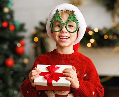 Smiling little boy wearing funny glasses in form of Christmas trees    holding Xmas gift while standing  in a decorated holiday room at home, dressed in warm red xmas sweater.