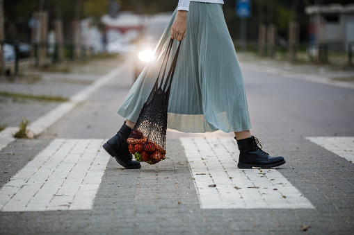 Low section of unrecognizable young woman, in a flowy skirt, carrying a reusable mesh bag with groceries and crossing the street on the crosswalk. Car headlights in the background.