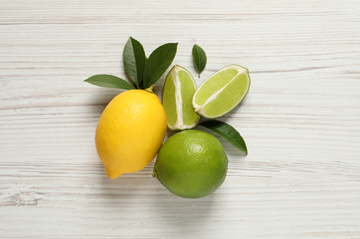 Fresh ripe lemon, limes and green leaves on white wooden background, flat lay