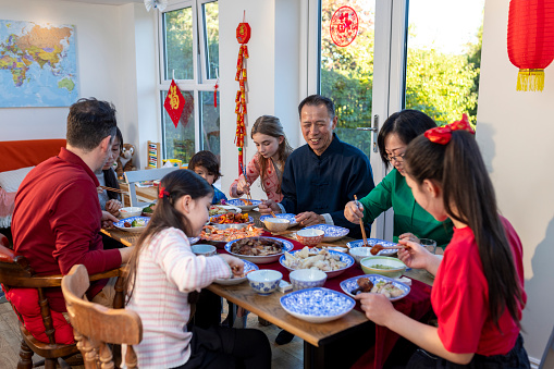 An extended family sitting at a dining table and eating a meal during Chinese New Year at home in Newcastle Upon Tyne, England. There are dishes with different Chinese food on them on the table for the family to choose from.