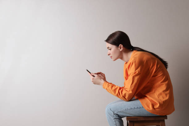 mulher nova com má postura usando o smartphone enquanto sentada no banquinho contra o fundo cinza claro, espaço para o texto - wrong side - fotografias e filmes do acervo