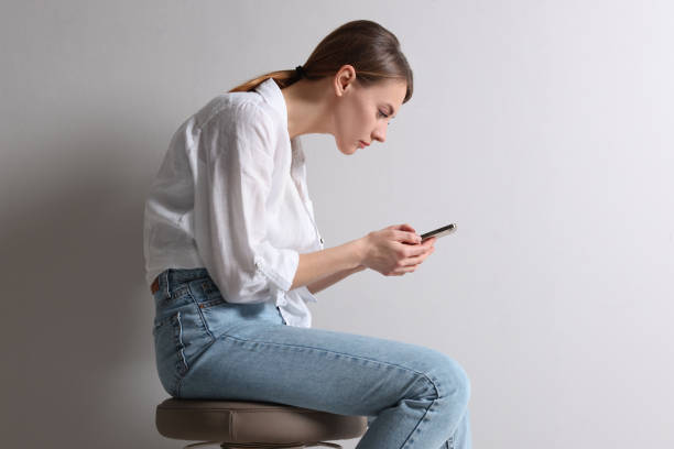 mujer con mala postura usando un teléfono inteligente mientras está sentada en un taburete sobre un fondo gris claro - posture fotografías e imágenes de stock