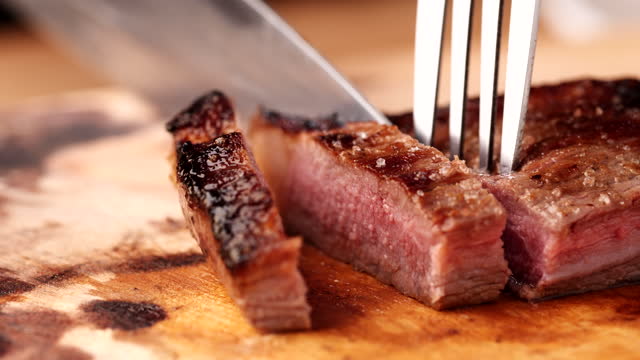 Chef slicing medium or well done beef steak with knife and fork on wooden cutting or chopping board, 4K. Concept of meat, beef steak