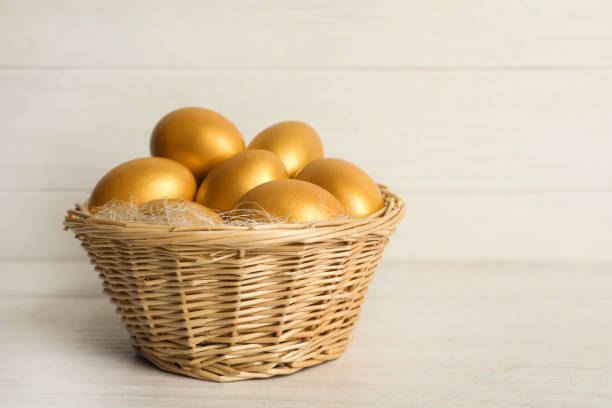 白い木のテーブルの上の枝編み細工品バスケットに光沢のある金色の卵、テキスト用のスペース - shiny group of objects gem bright ストックフォトと画像