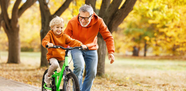 un grand-père de famille heureux apprend à son petit-fils à faire du vélo dans un parc - family bicycle cycling healthy lifestyle photos et images de collection