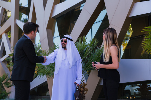 Young entrepreneur man with their colleagues presenting and shake hand with Emirati Arab Businessman about a new business projects and plans with digital tablet to poised and successful on behind of modern office outside. UAE