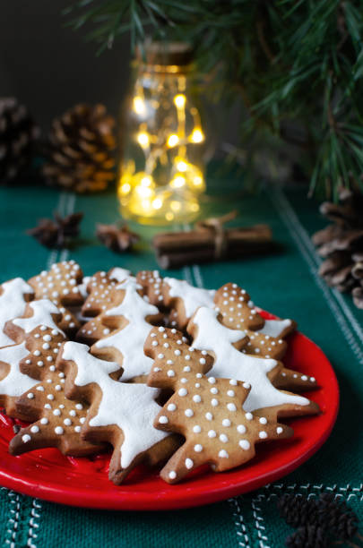 istock Festive cookies decorated with white icing on a red plate on a green background with lights. Homemade ginger and cinnamon Christmas cookies. Vertical orientation. Selective focus. 1445756865