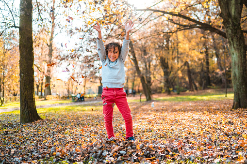 Full length shot front view of a young multiracial boy playing with autumn leaves in a public park. He has his arms up and a toothy smile. He is happy and cheerful.