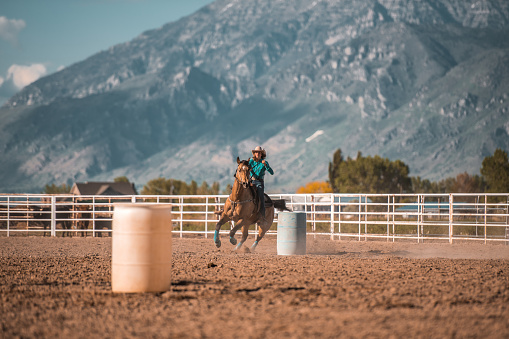 Young cowgirl is horseback riding and barrel racing in rodeo arena in Utah, USA. Sunlight in the back.