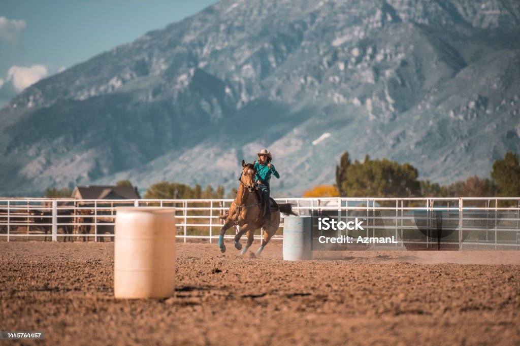 Young Cowgirl Barrel Racing With Horse Young cowgirl is horseback riding and barrel racing in rodeo arena in Utah, USA. Sunlight in the back. Rodeo Stock Photo