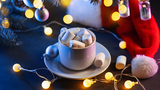 A cup of hot cocoa on the Christmas table with marshmallows sprinkled with cinnamon. Round vintage garland and Santa Claus hat.