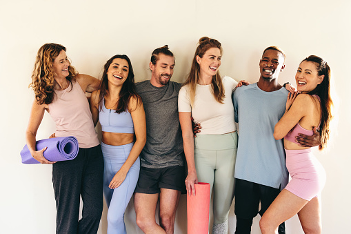 Diverse people laughing happily while standing against a wall with yoga mats. Multicultural people attending a yoga class together. Group of sporty people working out in a community yoga studio.