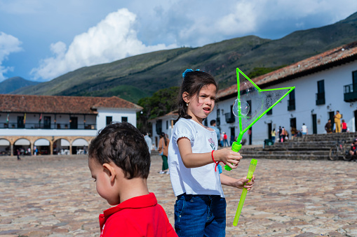 Latino brothers with an average age of 7 and 3 years old dressed casually are in the historic square of Villa de Leyva where they traveled on vacation with their family playing with a bubbler to make giant bubbles while running and having fun in the breeze