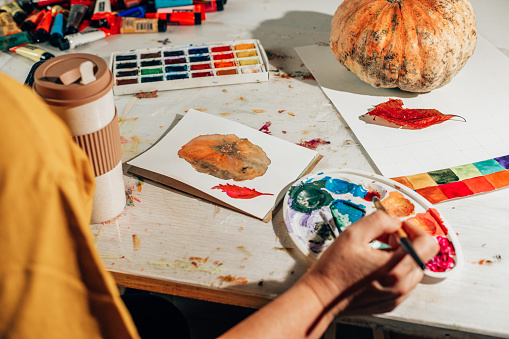 A woman paints a picture with a brush in a sketchbook. Learning to draw at home. Autumn still life with pumpkin and red leaf. Creating content concept.