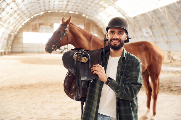 istock With saddle in hands. Young man with a horse is in the hangar 1445775877