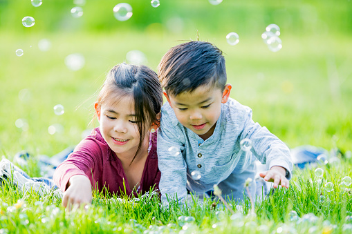 A sweet little brother and sister of Asian decent lay in the grass together as they blow bubbles in the wind on a warm spring day.  They are both dressed casually and are smiling.