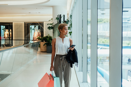Happy businesswoman typing text message on her smartphone and holding paper bags while standing in the modern shopping mall with big windows.
