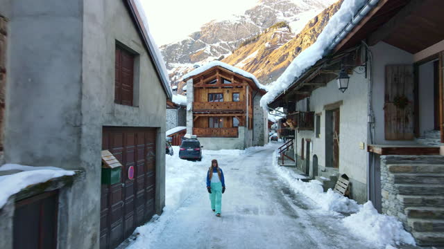 Picturesque small Swiss village at the foot of the rock Alps mountains, drone flying along the narrow street with frozen road and walking tourist woman, traditional cottages and chalets covered with snow. Alps tourism and ski resorts, winter in Switzerla