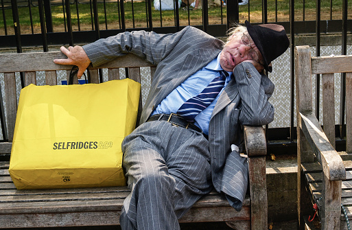 London England - July 3 2009; Elderly man well dressed asleep on street bench seat with bright yellow Selfridges shopping bag, London