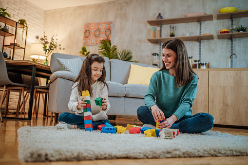 Mother and daughter playing together with colorful toys blocks on a carpet on the floor at home