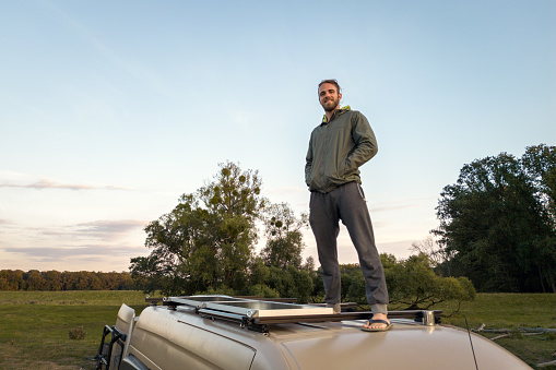 Man standing on the roof of a camper van parked in nature. Late summer evening