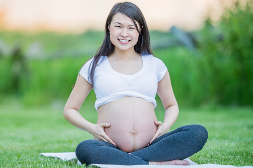 A young pregnant Asian woman sits outside on a yoga mat as she poses for a portrait.  She is dressed comfortably as she embraces her growing belly and sits peacefully.
