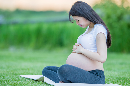 A young pregnant Asian woman sits outside on a yoga mat as she poses for a portrait.  She is dressed comfortably as she embraces her growing belly and gases at it in wonder.