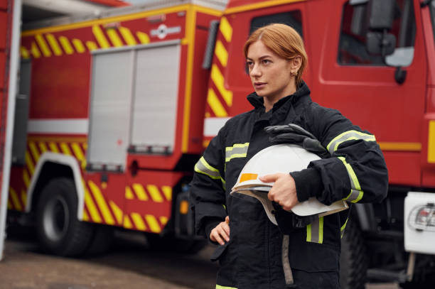 Heavy truck behind. Holding protective helmet. Woman firefighter in uniform is at work in department stock photo