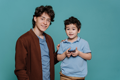 Two caucasian brothers isolated over turquoise studio backdrop smiling dressed in casual. Elder brother toothy smiles looks at camera, little boy holds phone. Brotherhood and family concept.