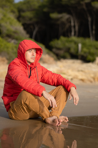 young adult man meditating on the sand on the shore of the beach. relaxing in front of the ocean. mindfulness and wellness concept.