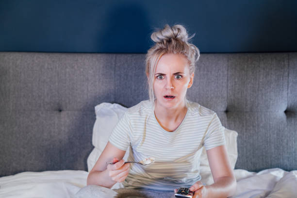 istock Shocked, stressed, surprised woman eating ice cream, holding a remote control while watching TV in bed. Scary horror movie or terrible news, scare in her eyes Overeating, Jamming of negative emotions. 1445803773