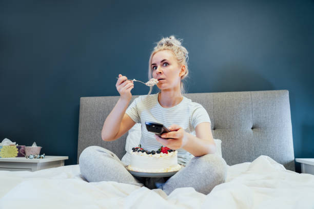 istock Surprised, stressed, shocked woman eating cake and holding a remote control while watching TV in bed. Scary horror movie or terrible news, scare in her eyes Overeating, Jamming of negative emotions 1445803776
