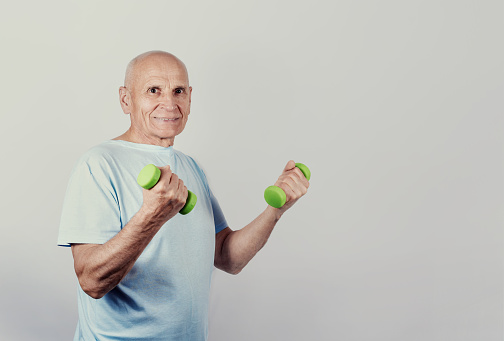 Sporty grandpa working with green dumbbells in hands in gym isolated on grey background. Healthy lifestyle concept.