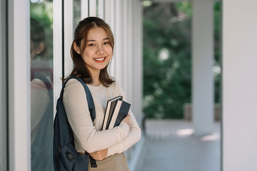 Cute Asian female student standing holding a book looking at the camera.