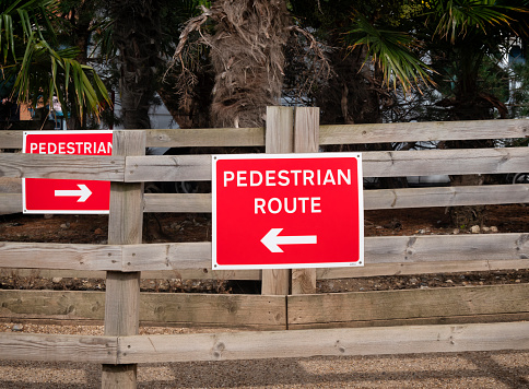 Two red “Pedestrian Route” signs on parallel fences, directing pedestrians to the left and to the right in separate lanes. Erected during the Covid pandemic, they remain in place to regulate the flow of tourists at this beach in Norfolk, Eastern England.