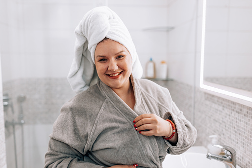 Adorable smiling plus size young Caucasian woman standing in bathroom after shower and smiling