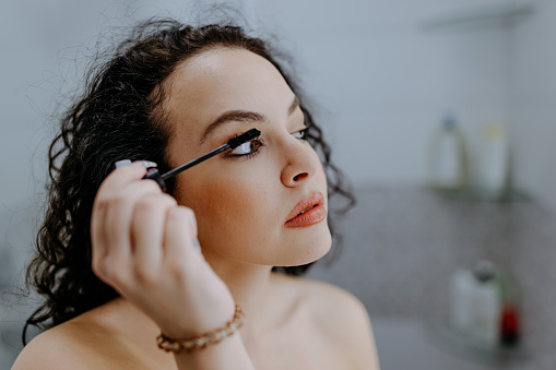 Close up beautiful focused young Caucasian woman applying some mascara in bathroom