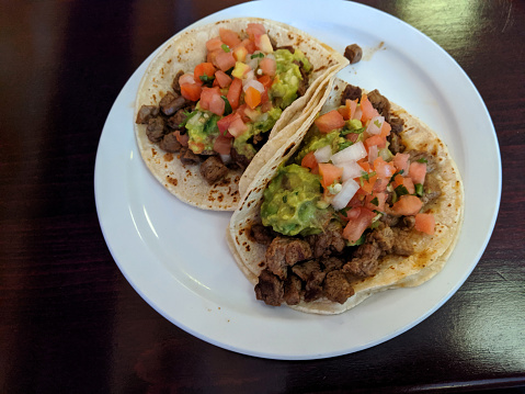 Two Steak Tacos topped with guacamole and pico de gallo on a white plate.