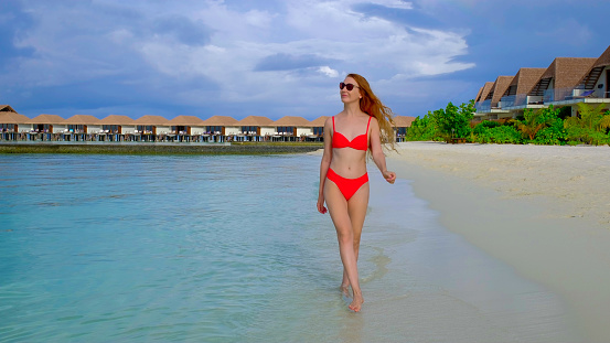 Woman in a bikini is walking. Female is happy on Maldives. Blue turquoise ocean on the background. Girl enjoys her tropical holidays. Summer travel vacation concept.