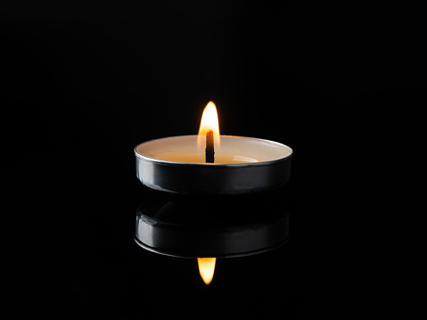 Burning tea light candle in the darkness. Lit candle is reflected in a black glossy surface. Focus on a single candlelight against black background. Fire in the night. Macro. Front view.
