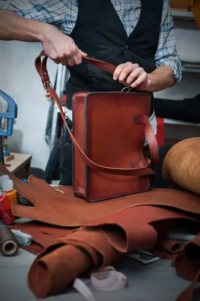 a craftsman in a workshop attaches a shoulder belt to a ready-made handmade leather briefcase