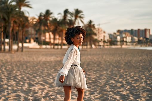 Cute African American child girl in a white dress with afro curls standing on a sandy beach by the sea against the backdrop of coastal houses.