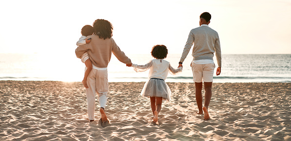 African American family with a cute curly daughter in a white dress and a baby son holding hands walking along the sandy beach near the sea at dawn.