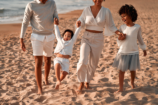 African American family with a cute daughter with afro curls in a white dress and a baby son walking and having fun holding hands on a sandy beach near the sea at dawn.
